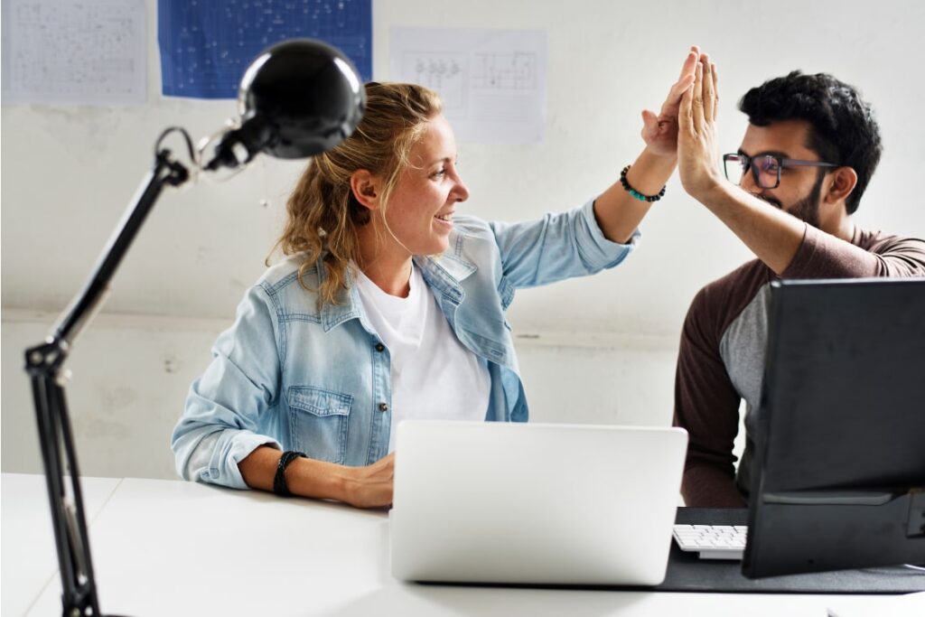 Female and male colleague high-fiving at a table with open laptops.