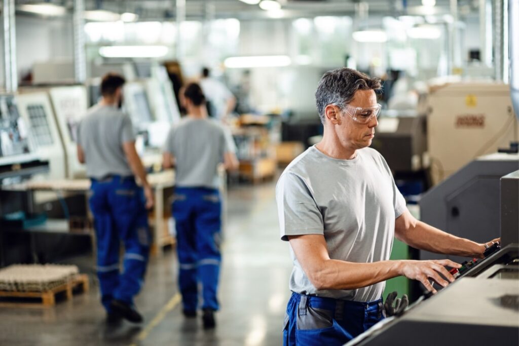 Male worker programming a CNC machine, with two colleagues discussing in the background near other machines.