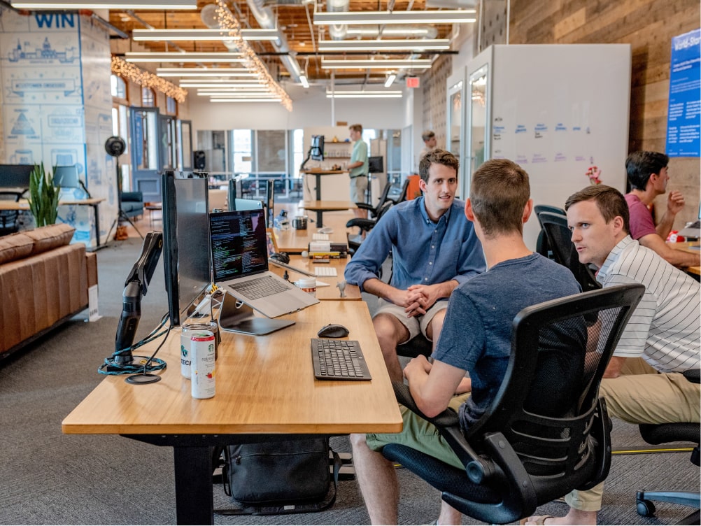 Three tech employees engaged in a discussion while looking at computer screens at a wooden table.